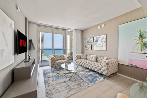 Modern living room with plush seating, glass coffee table, and floor-to-ceiling windows leading to a balcony. Enjoy bright spaces and ocean views from this inviting area designed for relaxation and socializing.