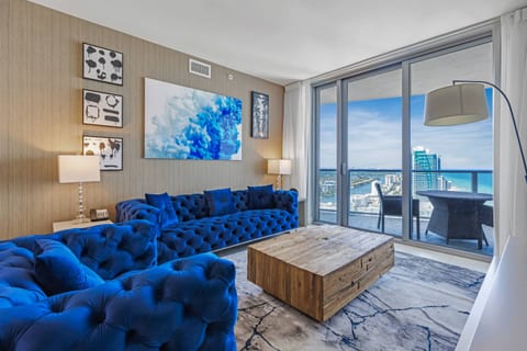 Step into your coastal retreat, featuring vibrant blue seating, a rustic coffee table, and expansive windows that open to sweeping balcony views of the ocean and city.