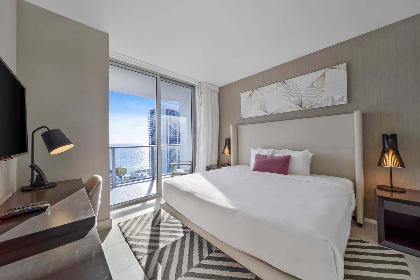 Step into the master bedroom featuring a king-size bed, modern lamps, and a work desk. Floor-to-ceiling windows open to a balcony with stunning ocean views. A private in-room bathroom adds convenience and comfort to this relaxing retreat.