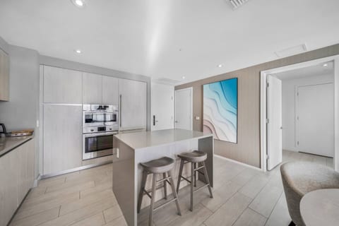 Sleek kitchen with wood cabinets, a spacious island with two stools, built-in stainless steel appliances, and a coffee maker. Open design flows into dining and living areas, ideal for easy entertaining.