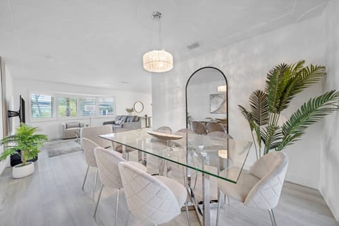 The dining area features a sleek glass table surrounded by eight cushioned chairs in soft beige tones. A stylish chandelier hangs above, creating an elegant space perfect for gatherings and meals.