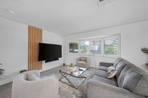 The living room features a comfortable gray sofa and two plush chairs around a glass coffee table. Large windows with shutters allow natural light to fill the room, creating a bright and welcoming space to unwind.