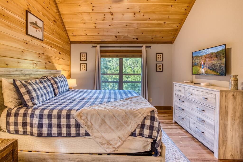 Vaulted ceilings above a grand king size bed. The main floor bedroom has views of the mountains and sky with en-suite bathroom attached. Flat screen television above dresser and charging station on nightstand for devices
