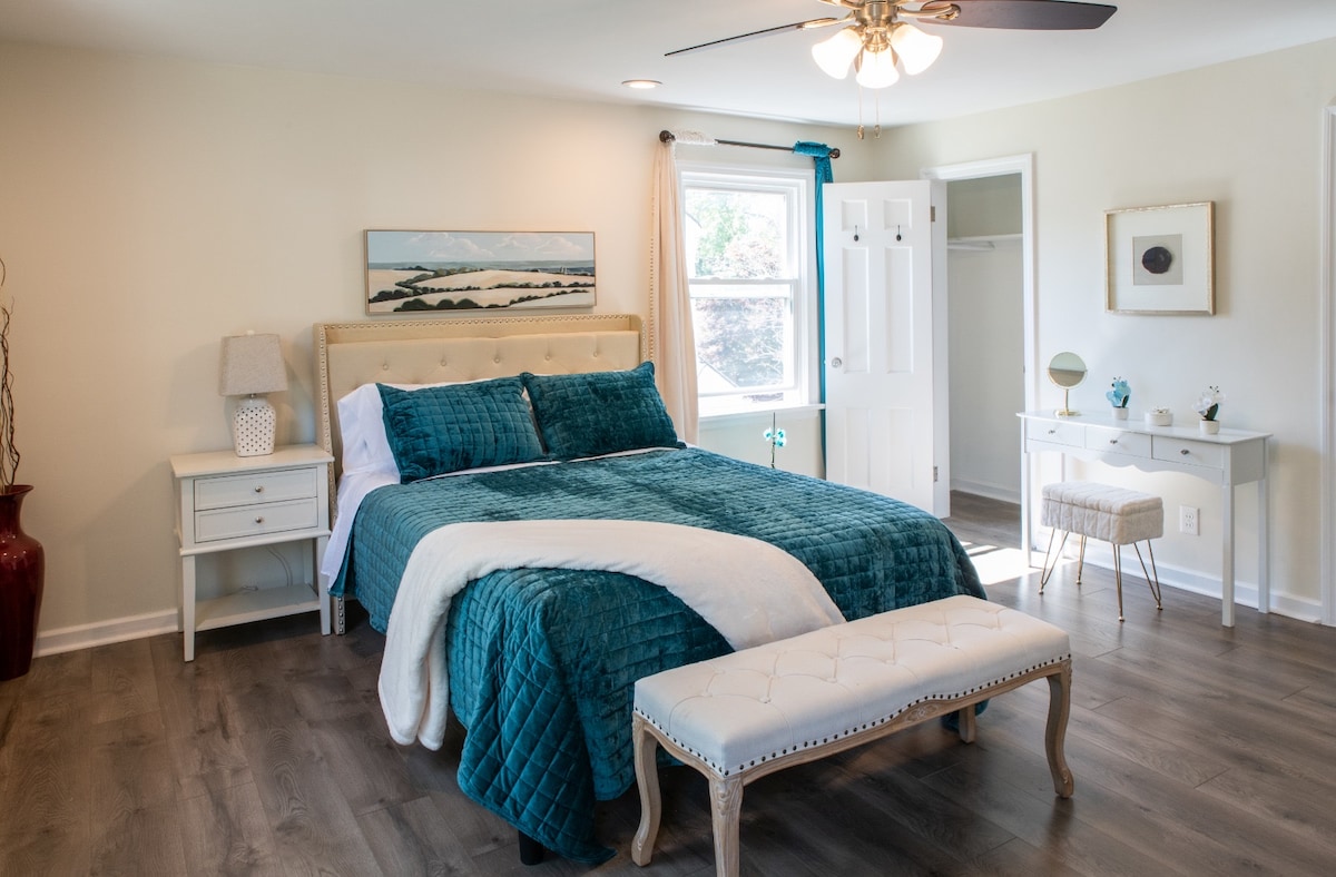 This bedroom has a light and airy feel with a touch of coastal charm. The use of neutral colors and natural light creates a relaxing and inviting space. The room features a queen-size bed and a teal duvet cover. and decorative pillows