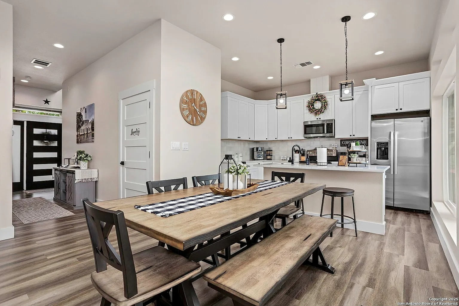 Dine in style at this chic wooden table with mixed seating, flowing into a bright white kitchen with stainless steel appliances, pendant lighting, and a cozy rustic touch. Perfect for meals & mingling!