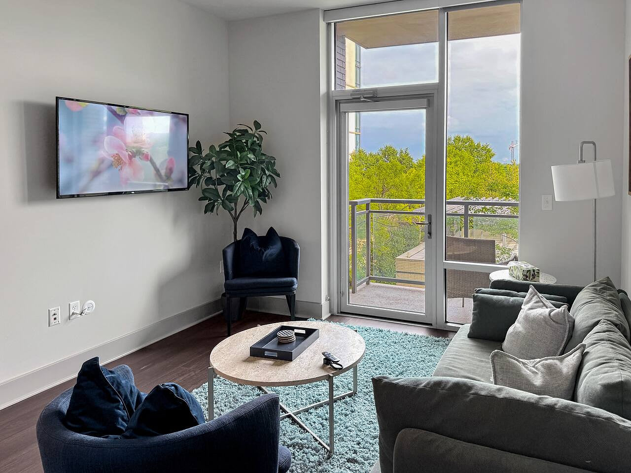 Bright, modern living room with cozy seating, a wall-mounted TV, and balcony views through floor-to-ceiling windows.