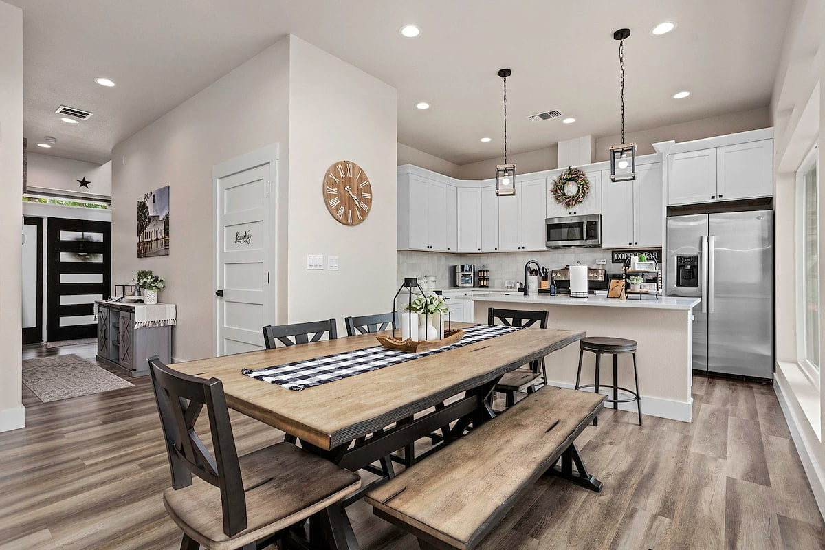 Dine in style at this chic wooden table with mixed seating, flowing into a bright white kitchen with stainless steel appliances, pendant lighting, and a cozy rustic touch. Perfect for meals & mingling!