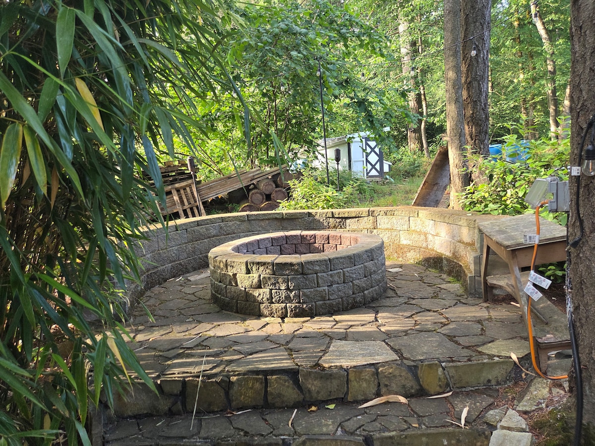 Flagstone patio with large outdoor firepit, surrounded by seating and backed by forest. Outdoor string lights available for added ambiance.