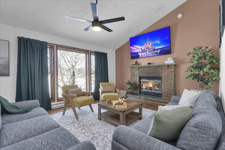 Spacious lakeview living room featuring two oversized couches, extra seating, a large TV, cozy fireplace, and ceiling fan—perfect for relaxing together after a day of fun.