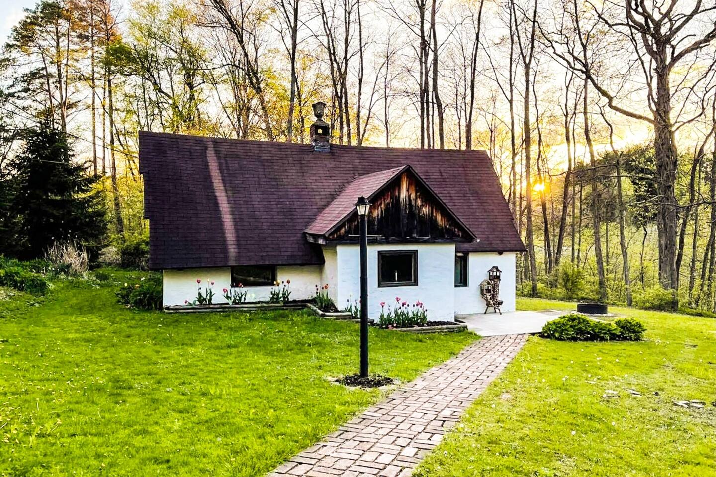 Escape to our charming country side cottage nestled in a verdant forest. Admire the cozy scenery, homely white facade, and a winding brick path leading to sunlit expanses. A tranquil retreat for nature lovers! You can see the lake through the trees!