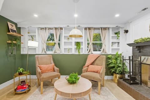 This cozy corner of the Green Hideaway includes chairs, a table, lamps, a plant, and the fireplace for a warm, inviting feel.