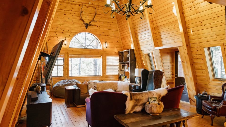 Spacious A-frame living room with dramatic vaulted ceilings, statement chandelier, and multiple seating areas—ideal for gathering, movie nights, and relaxing in a warm, rustic White Mountains cabin retreat.
