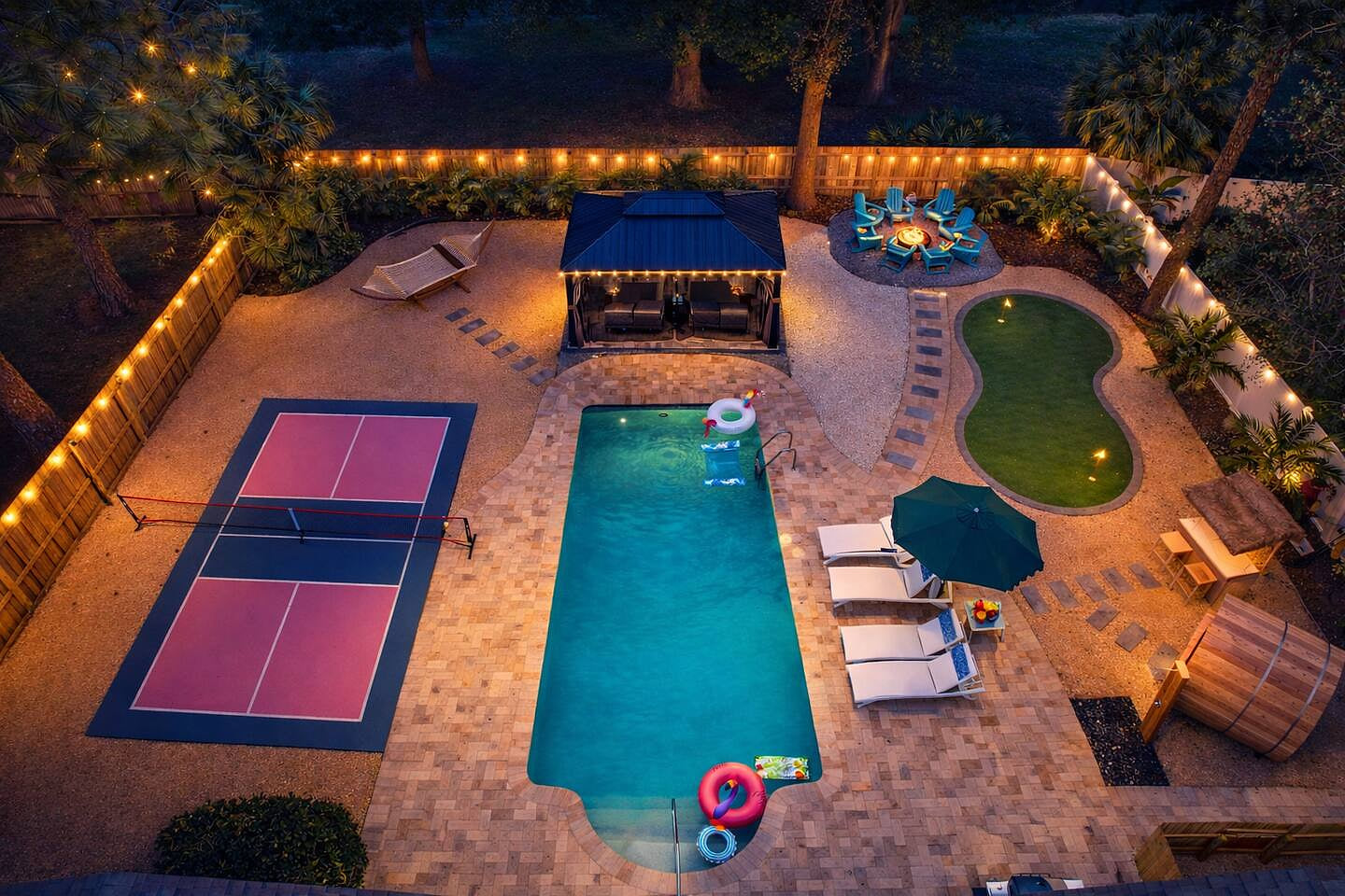 A private resort oasis after dark- heated pool glowing under café lights, pickleball court, fire pit lounge, cabana seating, and total privacy. Designed for unforgettable evenings and elevated beach living, just 5 minutes from the shore.