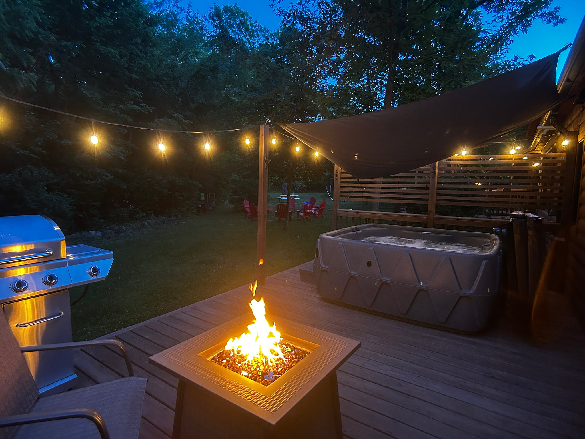 Private deck with fire table, seating, BBQ grill, hot tub, and glowing café lights—perfect for relaxing nights outdoors.