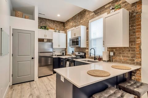 Stylish modern kitchen with exposed brick, quartz countertops, and stainless steel appliances. Fully equipped for cooking, with sleek cabinetry, gas stove, microwave, and breakfast bar—perfect for short or extended stays