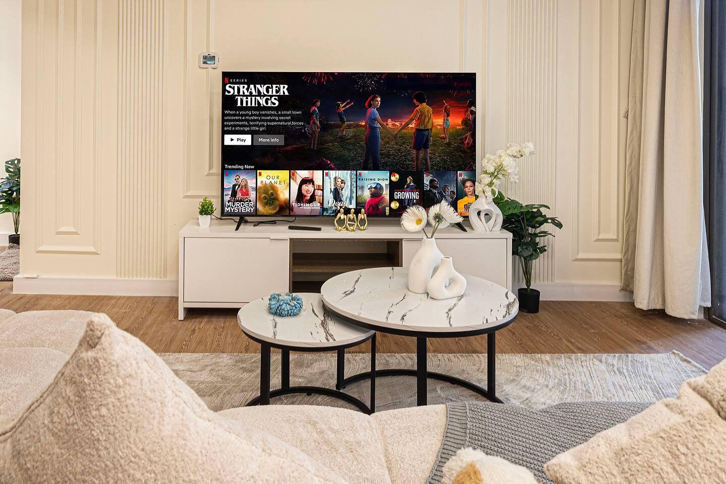 Modern living room with large TV showing "STRANGER THINGS", white console, two circular marble coffee tables, light sofa, and decorative plants.