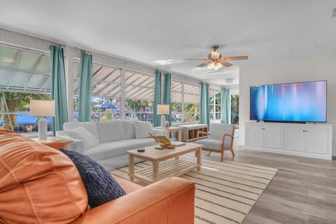 Sunlit coastal lounge with wall-to-wall windows, comfy seating, and a big smart TV