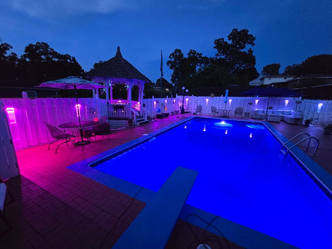 A stunning nighttime view of the private pool, glowing with vibrant LED lighting. The spacious patio features a lounge area with outdoor sofas, a dining table with an umbrella, and a charming gazebo in the background.
