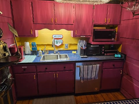 A vibrant, maximalist kitchen featuring bold magenta cabinets and a sunny yellow backsplash. Includes a blue tiled countertop, stainless steel sink, microwave, toaster oven, and coffee maker—perfect for a colorful morning start.