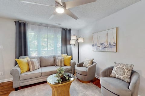 A bright, cozy living room with a gray sofa and yellow pillows, wicker coffee table with a plant, two armchairs, sailboat wall art, floor lamp, sheer curtains, hardwood floors, and a ceiling fan creating a calm, airy feel