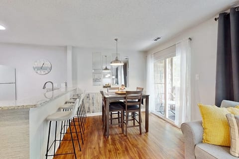 A dining table with chairs is set up in front of a wall with mirrors. To the left is a kitchen counter with bar stools.