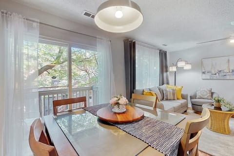 A dining area with a glass table is in the foreground, overlooking a living room with a sofa and armchair. A large window with a view of trees is to the left