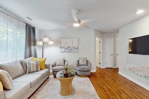 Bright living room with a sofa and two armchairs around a small wicker coffee table. Sheer curtains let in sunlight, a floor lamp and ceiling fan add light, and a wall-mounted TV, warm wood floors, and simple art create a cozy, inviting space.
