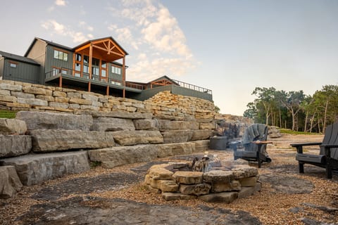 Stone fire pit with Adirondack chairs and a s'mores setup. Gather on the lower terrace as the evening cools, with the timber-gable house rising above.