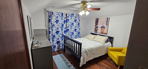 This bright and cozy bedroom features a comfortable full-size bed, bold patterned curtains, a ceiling fan, and a cheerful reading chair. A dresser and mirror add function and style—perfect for relaxing and recharging during your stay.