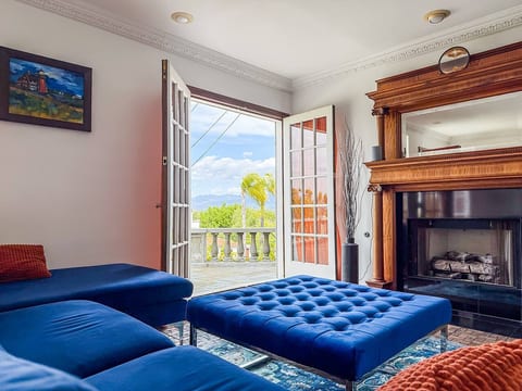 Family Room Retreat w/ a blue velvet sectional, tufted ottoman & ornate fireplace. French doors open to a sunlit terrace and treetop vistas - ideal for morning coffee or evening lounging.