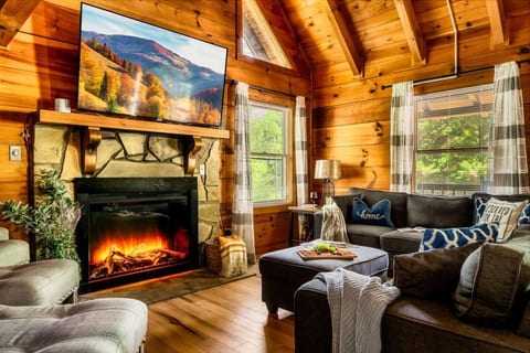 Cozy log cabin living room with stone fireplace, large flat-screen TV, vaulted wood ceiling, and plush seating for the whole group