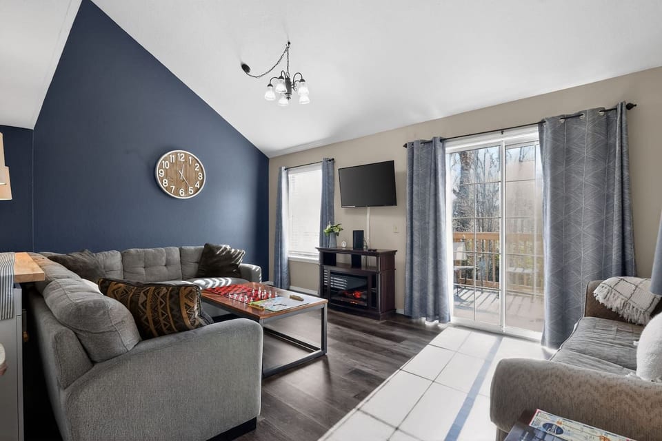 Cozy living room with two plush couches (one a sofa bed), throw blankets & pillows, fireplace, coffee table with games, Roku TV with Disney+ & Hulu, leading to master bedroom, bath, and twin bed upstairs.
