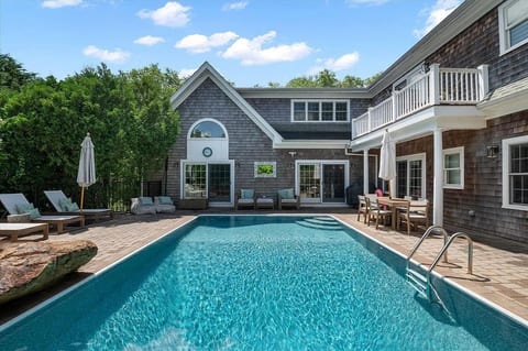 The classic Hamptons shingle exterior frames a sparkling saltwater pool, with the signature arched window of the living room visible above.