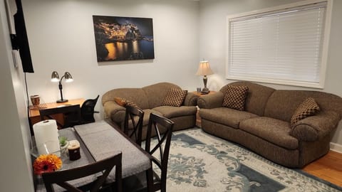 To the right: two plush couches and a large window. Opposite you: a mounted TV and a table with four chairs. To the right of the table is a corner desk with a lamp. A large rug anchors the center floor. Simple, clear, and easy to navigate.