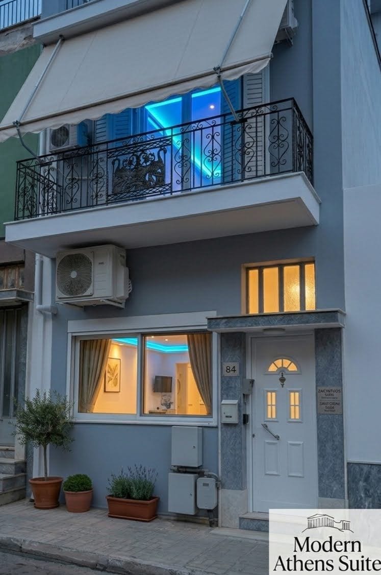 An exterior view of the Modern Athens Suite building, showcasing the ground-floor Zakynthos Suite and the first-floor Santorini Suite, both illuminated with their signature vibrant ambient lighting.