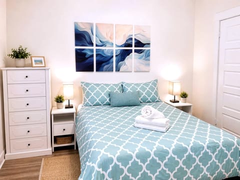 Bright coastal bedroom with queen bed, soft lighting, and fresh towels.