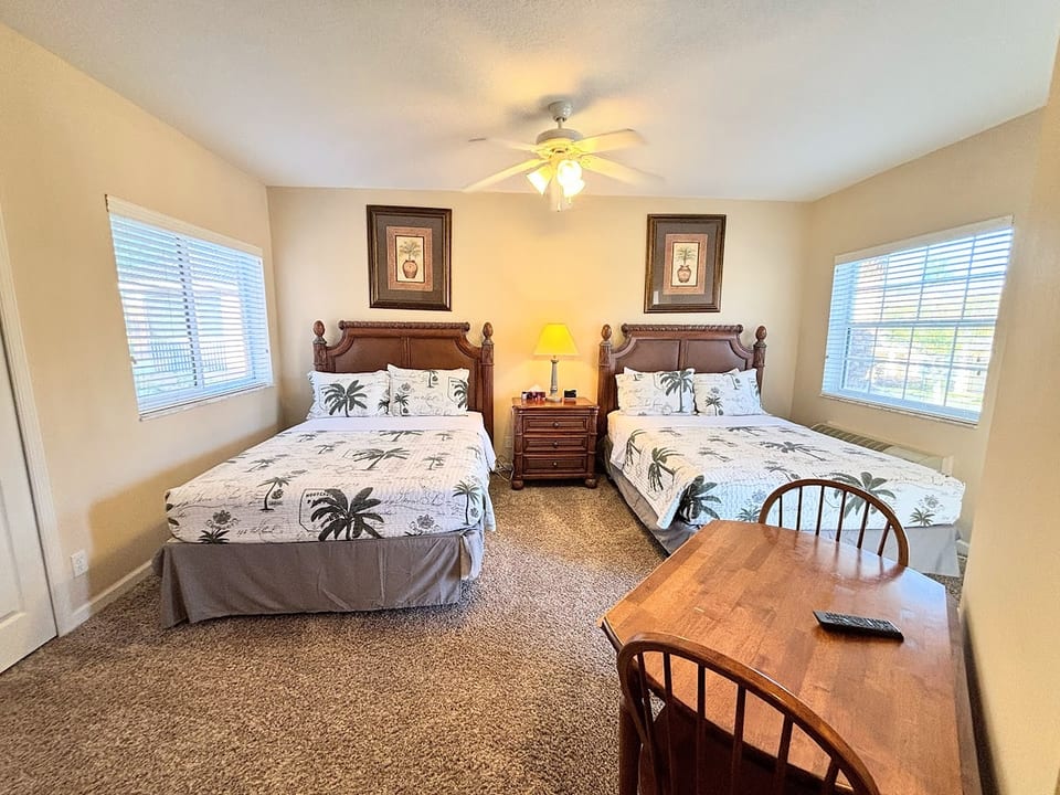 Spacious bedroom featuring two comfortable beds, ceiling fan, small dining/work table, and plenty of natural light.