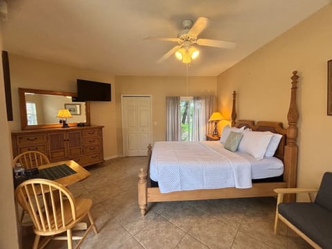 Comfortable bedroom with a large bed, wall-mounted TV, ceiling fan, small dining/work table, and a bright, relaxing layout.