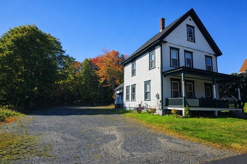 Historic farmhouse with covered porch, large driveway, and wooded surroundings with fall foliage.