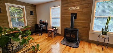 Cozy corner with a wood stove, workspace, large windows, and warm wood finishes throughout.