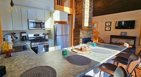 Open kitchen with bar seating, stainless steel appliances, and a stone fireplace overlooking the living area.
