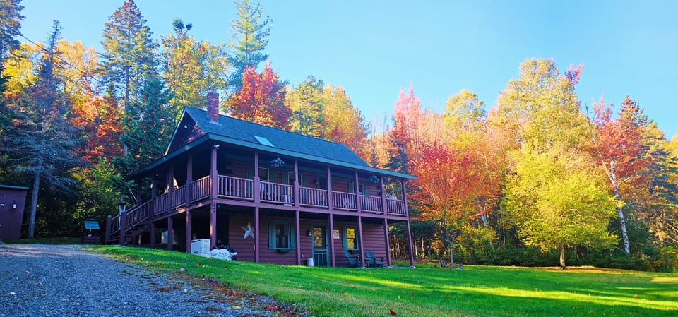Spacious log cabin surrounded by colorful fall foliage, featuring a wraparound porch and peaceful wooded setting.