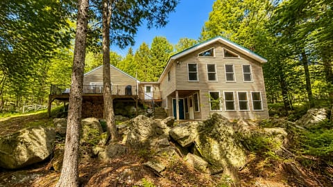 This spacious cabin is surrounded by trees and features a large deck, beautiful rock landscaping, and a peaceful woodland setting.