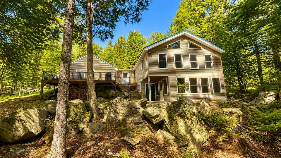 This spacious cabin is surrounded by trees and features a large deck, beautiful rock landscaping, and a peaceful woodland setting.