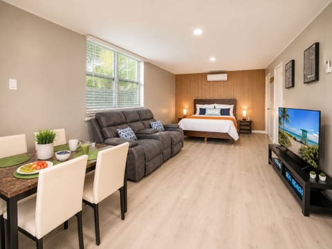 Wide studio overview featuring dining area, recliner sofa, king bed, wood-panel wall & large TV—everything in one stylish space.
