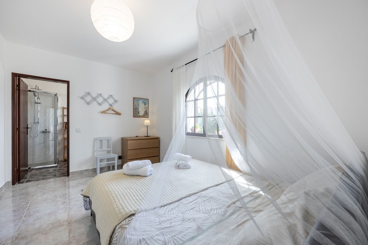 The bedroom features a 1.50m x 2m bed with a medium firmness mattress. The bathroom is accessible from the room. The window faces the shared garden area and has exterior shutters and light and dark curtains on the interior.