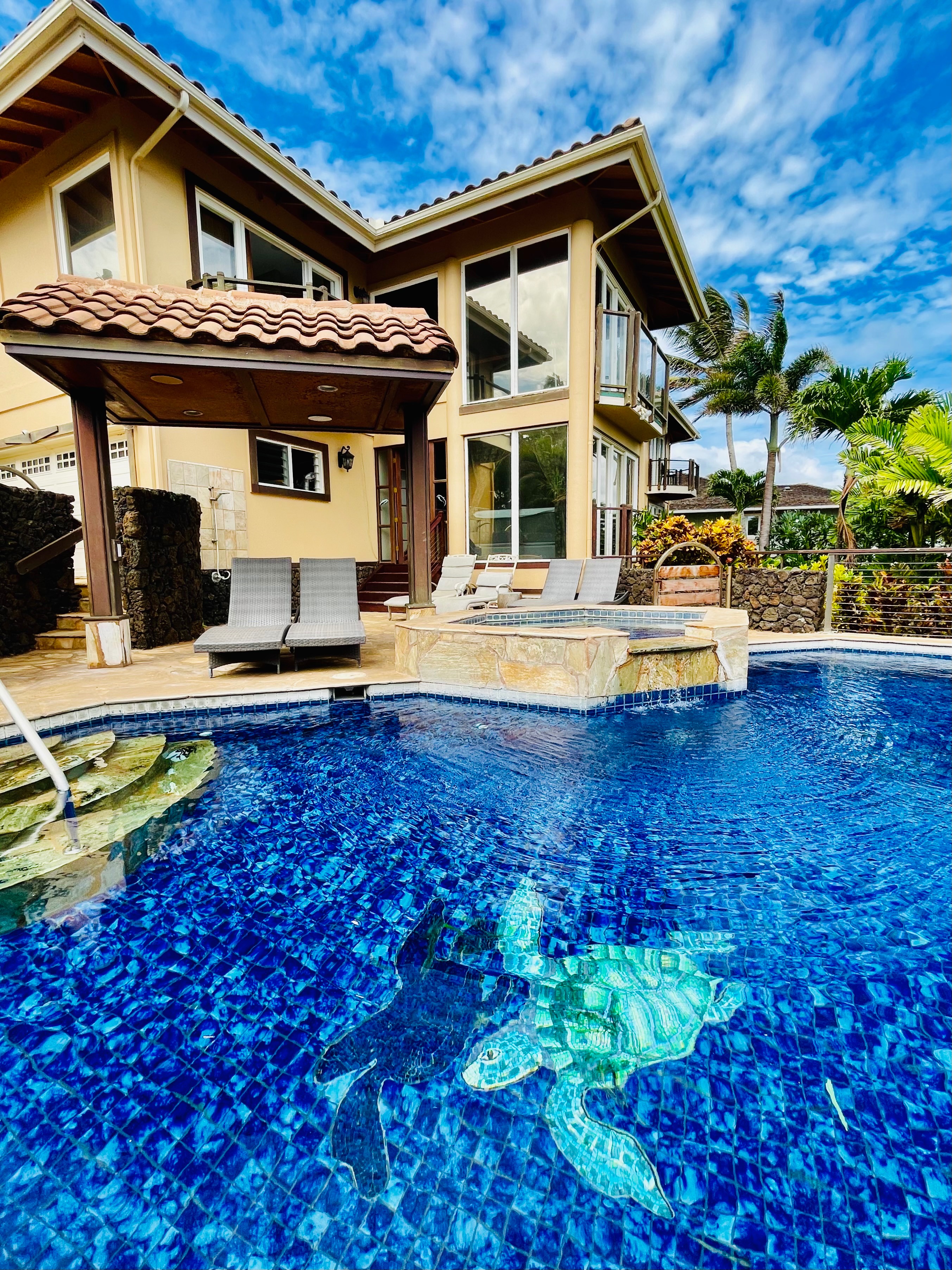 A luxurious two-story villa on Kauai’s South Shore featuring a stunning blue-tiled pool with mosaic sea turtles, surrounded by tropical palms and sun loungers. Expansive windows and island breezes create the perfect blend of elegance and paradise