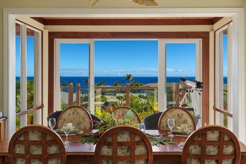 Elegant dining table set before wide glass doors, framing palm trees and a deep blue ocean. Warm wood accents, tropical-patterned chairs, and bright daylight create a relaxed, resort-style atmosphere