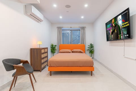 Bright, spacious bedroom with a bold orange statement bed, smart TV, and A/C for year-round comfort. Clean modern design, warm lighting, and a large window with soft curtains—your calm, boutique-style reset after the city