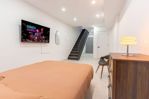 Wide-open, modern suite with smart TV, warm designer lighting, and a dedicated work nook—plus sleek stairs leading to the upper level. Clean lines, bright ceilings, and plenty of space to spread out and settle in